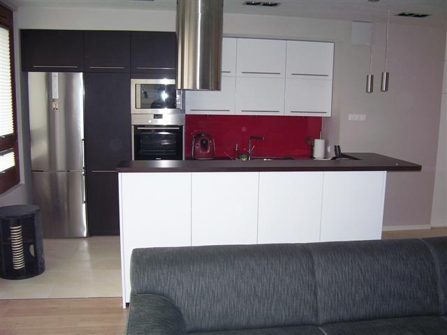 Nowoczesna kuchnia z czarnymi i białymi szafkami, stalowym sprzętem AGD i czerwonym panelem nad blatem, widziana z perspektywy salonu, z fragmentem szarej sofy w kadrze.